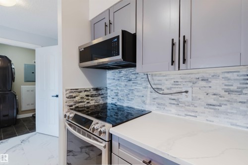 The kitchen features stainless steel appliances, grey cabinetry with black hardware, a tiled backsplash, and light-colored countertops - #204 11803 22 Av Sw, Edmonton, AB 
