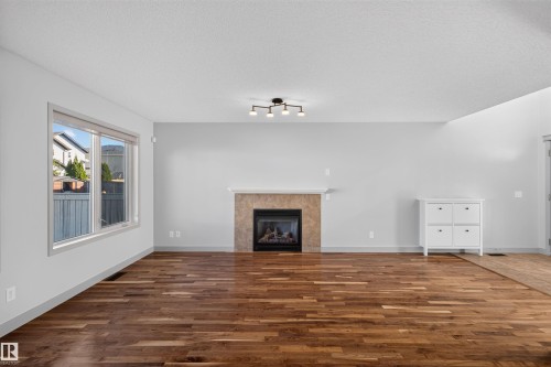 Wood-finish flooring, fireplace with a tiled surround, window with white trim, white walls, and a modern ceiling light fixture - 6116 8 Avenue Sw, Edmonton, AB 
