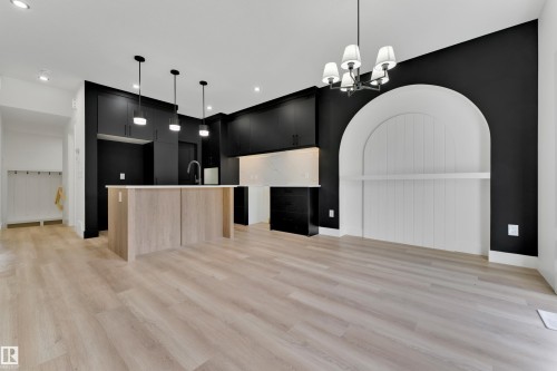 Open-concept living space with light wood-finish flooring, a kitchen island featuring light wood-finish paneling, dark cabinetry, and an arched built-in niche with a shelf - 261 Linden Loop, Leduc, AB 