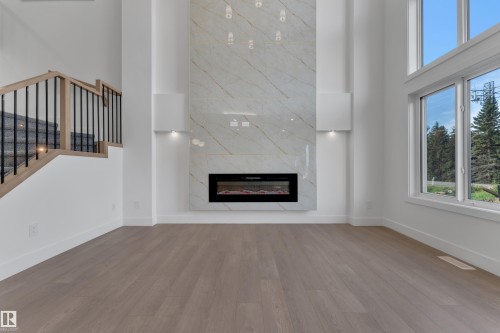Wood-finish flooring throughout, a modern electric fireplace with a white stone-look surround, a staircase with wood handrails and black metal balusters, and expansive windows - 247 Linden Loop, Leduc, AB 