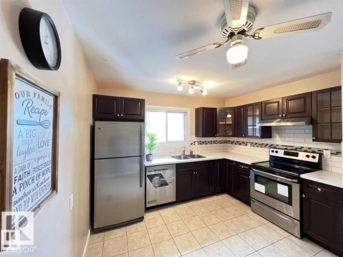 Kitchen featuring dark wood-finish cabinetry, stainless steel appliances, white subway tile backsplash, light-toned tiled flooring, and a ceiling fan with integrated lighting - 144 Kaskitayo Court Nw, Edmonton, AB 