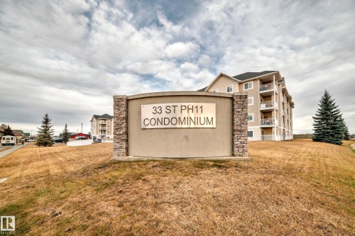Stone-clad monument sign with "33 ST PH11 CONDOMINIUM" inscription, set against a backdrop of a multi-story building featuring balconies and a dark-toned roof - #302 4309 33 St, Stony Plain, AB 
