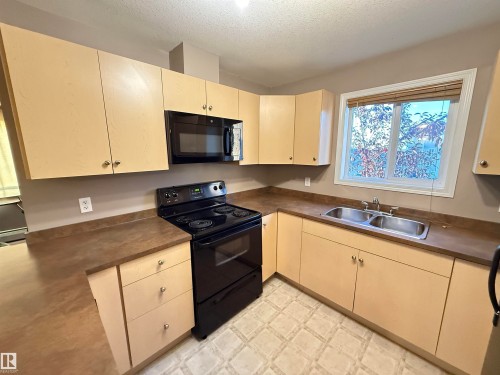 Light wood-finish cabinetry with silver-tone hardware, dark countertop surfaces, black range, and dual stainless steel sink - #302 4309 33 St, Stony Plain, AB 