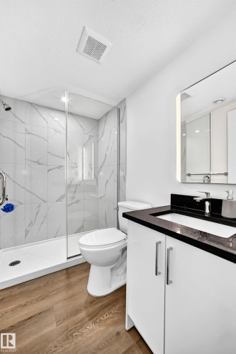 Contemporary bathroom featuring wood-finish flooring, a white vanity with chrome hardware and black countertop, and a walk-in shower with clear glass enclosure and marble-patterned tile surround - 1673 Cunningham Way Sw, Edmonton, AB 