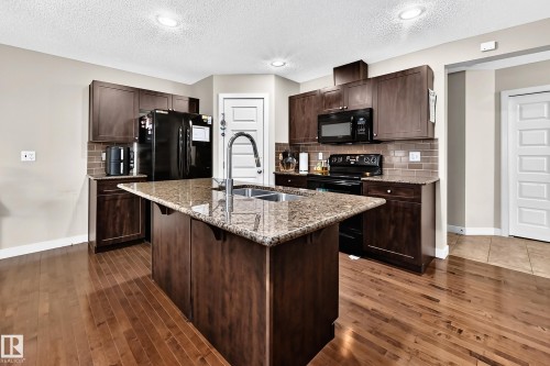 Kitchen featuring wood-finish flooring, dark wood cabinetry, a granite-finish island with an undermount sink, and black appliances - 1673 Cunningham Way Sw, Edmonton, AB 