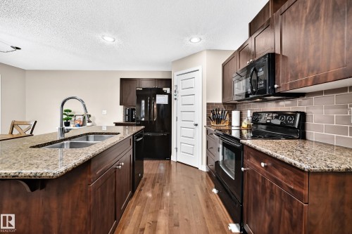 Kitchen featuring a central island with a double basin sink, granite countertops, dark wood-finish cabinetry, a black appliance suite, and a subway tile backsplash - 1673 Cunningham Way Sw, Edmonton, AB 