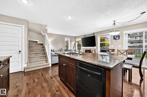 Open-concept living space with wood-finish flooring, a kitchen island featuring a built-in sink and dishwasher, and a staircase with carpeted treads - 1673 Cunningham Way Sw, Edmonton, AB 