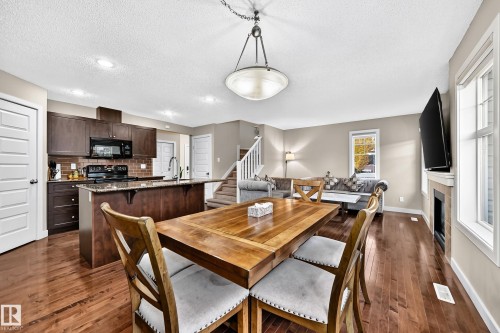 Open-concept living area featuring rich wood-finish flooring, a kitchen with dark cabinetry and stainless steel appliances, a central island, and a fireplace - 1673 Cunningham Way Sw, Edmonton, AB 