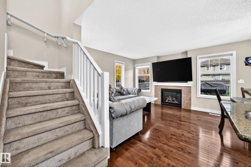 Spacious living area featuring rich wood-finish flooring, a contemporary fireplace with a stone surround, and large windows - 1673 Cunningham Way Sw, Edmonton, AB 