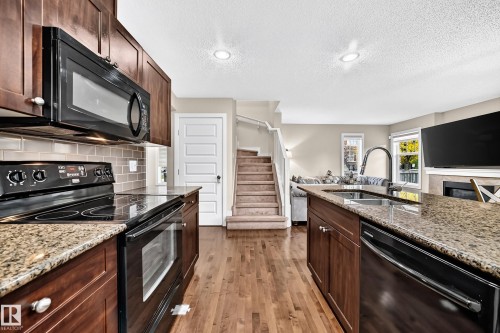 Wood-finish flooring throughout, dark wood-finish cabinetry, light-toned subway tile backsplash, recessed lighting, and a kitchen island with a double basin sink - 1673 Cunningham Way Sw, Edmonton, AB 