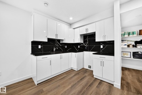 Modern white cabinetry with sleek hardware, black countertops, black marble-patterned backsplash, wood-finish flooring, and recessed lighting - 1673 Cunningham Way Sw, Edmonton, AB 