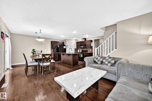 Open-concept living space featuring wood-finish flooring, a kitchen island with seating, dark wood cabinetry, and a staircase with white railings - 1673 Cunningham Way Sw, Edmonton, AB 