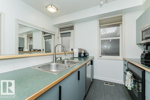 Kitchen featuring a double basin stainless steel sink with a high-arch faucet, gray cabinetry, built-in stainless steel appliances, and dark-toned floor tiling - #202 9503 101 Av Nw, Edmonton, AB 