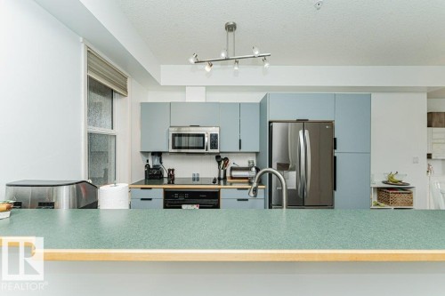 Well-appointed kitchen featuring light blue cabinetry, stainless steel appliances, a modern track lighting fixture, a stainless steel sink with a gooseneck faucet, and a breakfast bar with a speckled green countertop - #202 9503 101 Av Nw, Edmonton, AB 