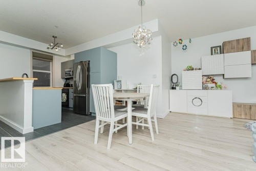 Open-concept living area with light wood-finish flooring, a contemporary chandelier, recessed lighting, and a kitchen featuring stainless steel appliances and a breakfast bar with a wood-finish countertop - #202 9503 101 Av Nw, Edmonton, AB 