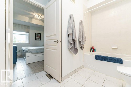 White tiled flooring leading to a bedroom with wood-finish flooring, a window with blinds, and a built-in ceiling light fixture - #202 9503 101 Av Nw, Edmonton, AB 