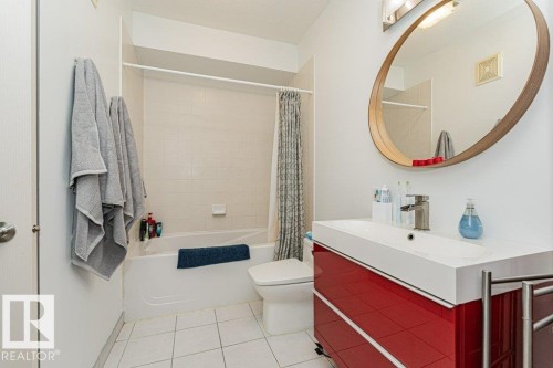White tiled flooring, white bathtub, white toilet, red vanity with integrated sink, and an oval mirror - #202 9503 101 Av Nw, Edmonton, AB 