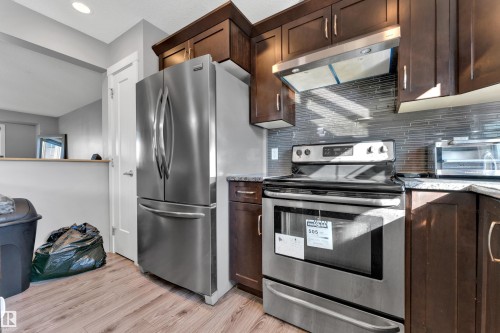 Contemporary kitchen featuring wood-finish flooring, stainless steel appliances, dark wood cabinetry, granite-style countertops, and a tiled backsplash - #22 4205 30 St Nw, Edmonton, AB 