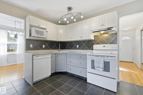 Kitchen featuring white upper cabinetry, grey lower cabinetry, tiled backsplash, white appliances, and a corner sink - 74 Aspen Crescent, St. Albert, AB 