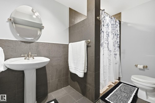 Pedestal sink with brushed nickel faucet, round mirror, dark grey tile wainscoting, and tiled shower area - 74 Aspen Crescent, St. Albert, AB 