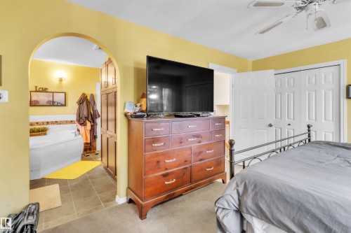 Primary Bedroom Suite with Ensuite Bathroom, Soft Yellow Walls, Wood-Finish Dresser with Television Mount, Ceiling Fan, White Panel Closet Doors, and Tile Flooring Throughout - 76 Gariepy Crescent Nw, Edmonton, AB 