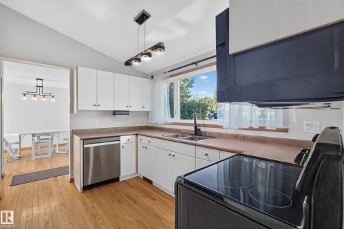 Kitchen featuring wood-finish flooring, white cabinetry, integrated stainless steel dishwasher, double basin sink with a black faucet, and a black electric range - 23 Clearwater, Devon, AB 