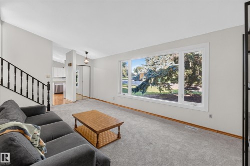 Expansive main-level room featuring a large picture window, light gray carpeting, and a staircase with a black metal railing - 23 Clearwater, Devon, AB 