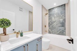 White soaking tub with a tiled accent wall, light blue vanity with gold hardware, gold-finish faucet, and a large frameless mirror - 