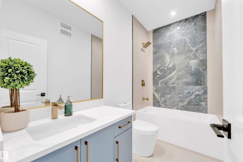 White soaking tub with a tiled accent wall, light blue vanity with gold hardware, gold-finish faucet, and a large frameless mirror - 2 Tenuto Link, Spruce Grove, AB 