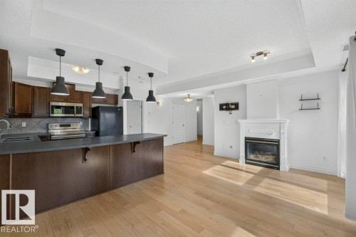 Open-concept layout featuring wood-finish flooring, a kitchen island with pendant lighting, dark wood cabinetry, and a built-in fireplace with a white mantel - #306 10232 115 St Nw, Edmonton, AB 