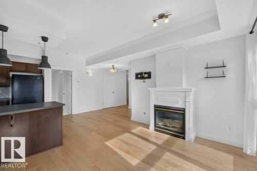 Wood-finish flooring, built-in fireplace with white mantel, tray ceiling, recessed lighting, and kitchen island with pendant lighting - #306 10232 115 St Nw, Edmonton, AB 