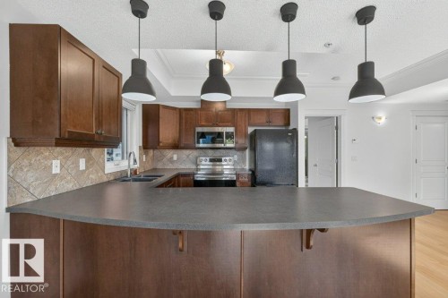 Kitchen featuring dark wood-finish cabinetry, a breakfast bar with a dark countertop, a tile backsplash, and stainless steel appliances - #306 10232 115 St Nw, Edmonton, AB 