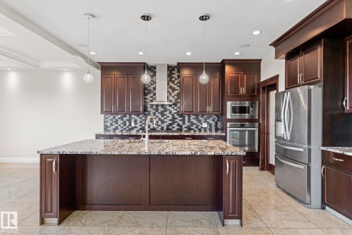 Spacious kitchen featuring an expansive center island with stone-finish countertop, dark wood-finish cabinetry, stainless steel appliances, and a mosaic tile backsplash - 229 Albany Drive Nw, Edmonton, AB 