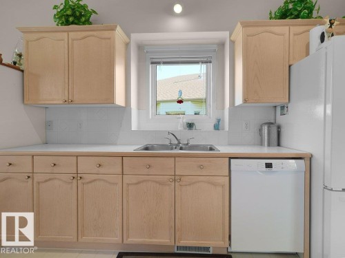Light wood-finish cabinetry, white countertops, stainless steel double-basin sink, tiled backsplash, and an integrated white dishwasher - 156 Phillips Row Nw, Edmonton, AB 