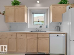 Light wood-finish cabinetry, white countertops, stainless steel double-basin sink, tiled backsplash, and an integrated white dishwasher - 