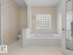 Built-in soaking tub, privacy glass block window, patterned wallpaper, toilet with grab bar, and light-colored flooring - 