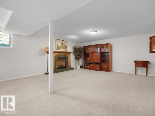 Expansive carpeted room featuring a fireplace with a wood mantel and green surround, a built-in wood-finish cabinet system, and a window with horizontal blinds - 156 Phillips Row Nw, Edmonton, AB 