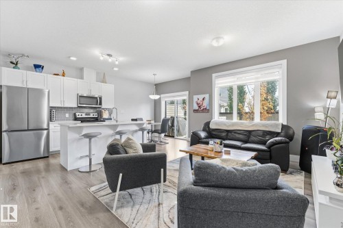 Open-concept living space with wood-finish flooring, a white kitchen island with counter seating, stainless steel appliances, and a sliding glass door - #15 4835 Wright Dr Sw, Edmonton, AB 