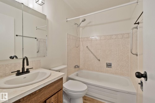 Bathroom featuring a single vanity with a white basin and matte black faucet, wood-finish cabinetry, a mirrored medicine cabinet, and a bathtub with a shower head and patterned tile surround - 14411 23 Street Nw, Edmonton, AB 