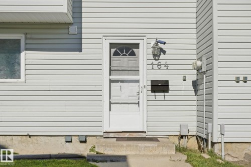 White vinyl siding exterior with an entry door featuring a storm door, an exterior wall lantern, a wall-mounted mailbox, and concrete steps - 164 Dickinsfield Court Nw, Edmonton, AB 