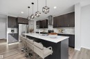 Contemporary kitchen featuring dark wood-finish cabinetry, stainless steel appliances, a white countertop island with seating, geometric pendant lighting, and light wood-finish flooring - 9227 223 Street Nw, Edmonton, AB 