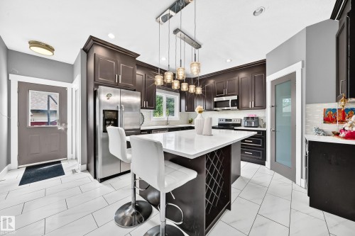 Spacious kitchen featuring dark wood cabinetry, stainless steel appliances, a large island with seating, pendant lighting, and light-toned tile flooring - 12730 118 Street Nw, Edmonton, AB 