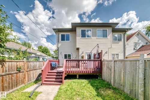 Rear exterior featuring light-colored siding, multiple windows, a red-stained wood deck with stairs, a concrete path, and a wood fence - 12730 118 Street Nw, Edmonton, AB 