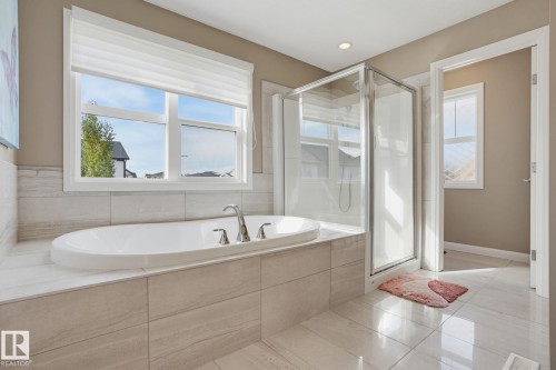Deep soaking tub with polished chrome fixtures, glass-enclosed shower, large window with blinds, and light-toned tile flooring - 9448 228 Street Nw, Edmonton, AB 