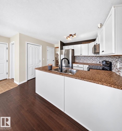 Kitchen featuring white cabinetry, a dual-basin stainless steel sink, dark wood-finish flooring, and a tiled backsplash - 447 Songhurst Place, Leduc, AB 