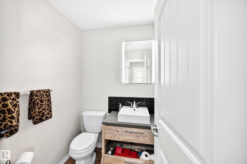 White paneled door opening to a bathroom featuring a rectangular vessel sink, dark stone countertop, wood-finish vanity with open shelving, and square wall-mounted mirror - 447 Songhurst Place, Leduc, AB 
