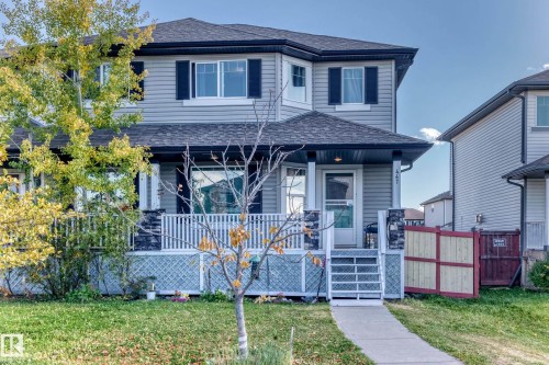 Two-story residence featuring light gray siding with contrasting dark shutters, a covered front porch with stone-clad pillars, and a peaked roofline - 447 Songhurst Place, Leduc, AB 