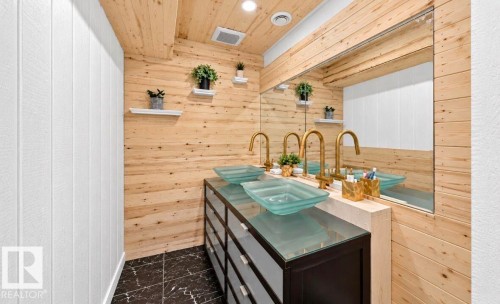 Dual vessel sinks with gold-finish faucets, black vanity with glass top, full-width mirror, wood paneling, and dark tile flooring - 447 Songhurst Place, Leduc, AB 