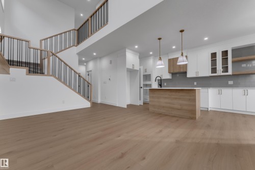 Open-concept living space featuring wood-finish flooring, a contemporary kitchen with white cabinetry, a large kitchen island, and a prominent staircase with dark metal balusters - 178 Munn Way, Leduc, AB 