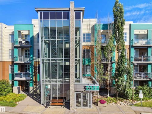 Modern multi-story building featuring a glass and metal facade, stone accents, multiple private balconies, and a prominent glass-enclosed stairwell - #415 2588 Anderson Wy Sw, Edmonton, AB 
